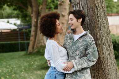 A female soldier in camouflage reunites with her wife, sharing a heartfelt embrace beneath a tree. clipart