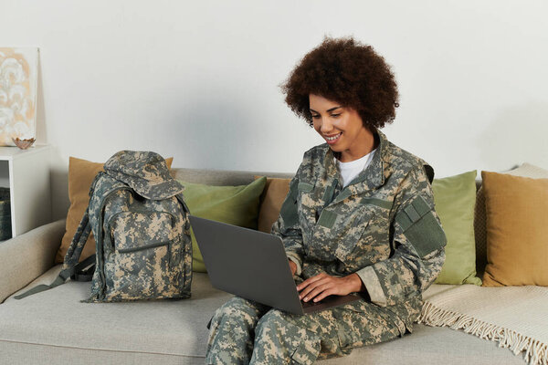 A young African American military woman in camouflage engages with her laptop in a cozy living space.