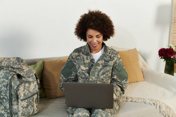 A female soldier in camouflage smiles while using her laptop on a cozy sofa in a military setting.