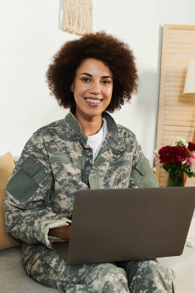 A confident young woman in camouflage uniform works on her laptop, smiling warmly in a cozy, bright setting.