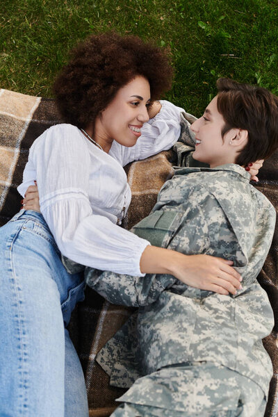 A soldier in camouflage reunites with her wife, sharing a tender moment on a cozy blanket.