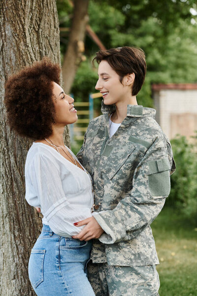 A young female soldier in camouflage shares a heartfelt reunion with her wife in a vibrant outdoor setting.