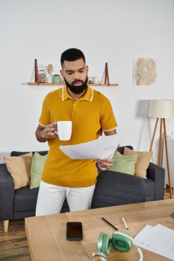 Man relaxes with a cup of coffee, focusing on papers in a stylish living space. clipart