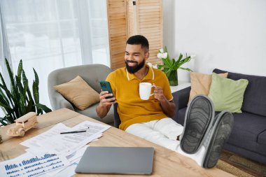 Neşeli bir adam evinde dinleniyor, kahvenin tadını çıkarıyor ve telefonunu karıştırıyor..