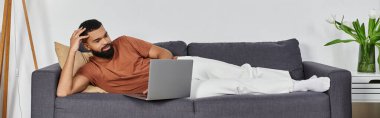 Handsome man lounges comfortably on the couch while browsing on his laptop. clipart