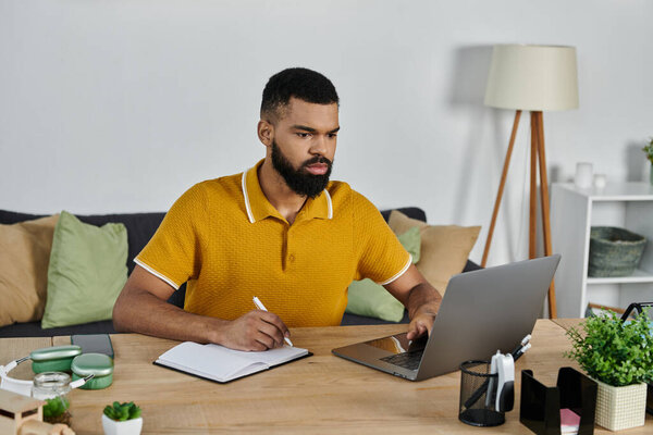 Focused individual writes notes while working on a laptop at home.