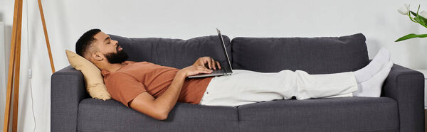 Красивый мужчина комфортно расслабляется дома, работая над ноутбуком на диване..