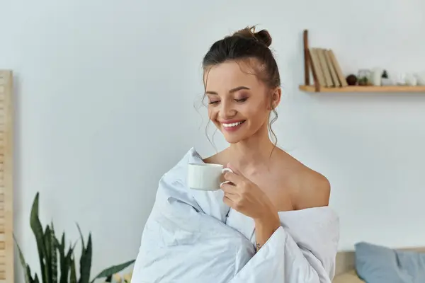 stock image A smiling woman sips coffee while wrapped in soft fabric in a serene setting.