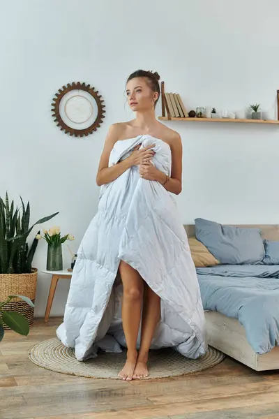 stock image A young woman stands elegantly, holding a blanket in a warmly decorated room.