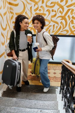 A cheerful couple stands together at a hostel, ready for their next adventure with bags and warm drinks. clipart