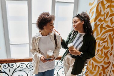 Two women share a joyful moment in a vibrant hostel, enjoying coffee and each others company. clipart