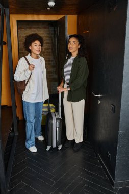 A happy couple stands by the door of their hostel room, excited for their travel adventure ahead. clipart