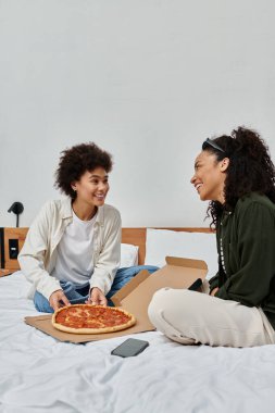 A happy couple shares laughs and delicious pizza while relaxing in their hotel room. clipart