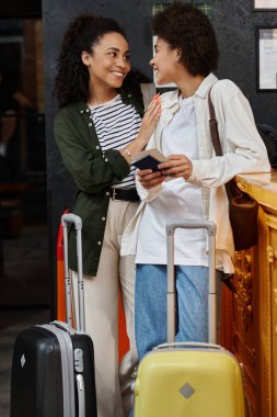 Two joyful women at a hostel reception, sharing smiles while checking in with their luggage. clipart