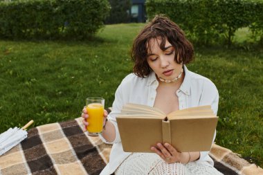 Beyaz gömlekli genç bir kadın piknik battaniyesinde rahatlıyor, kitap okurken içkinin tadını çıkarıyor..