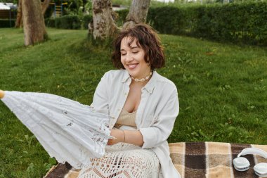 Beyaz gömlekli neşeli bir kadın yaz pikniğinde keyifli, sıcak bir gökyüzünün altında büyüleyici bir şemsiyeyle güneşleniyor..