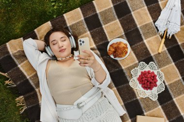 Beyaz gömlekli genç bir kadın çimenlerde dinleniyor, müzik ve lezzetli atıştırmalıklarla pikniğin tadını çıkarıyor..