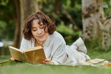 A young woman lounges on a blanket, immersed in a book, basking in the warmth of a summer day. clipart