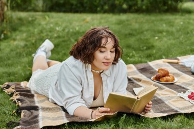 Beyaz gömlekli genç bir kadın, güneşli bir yaz pikniğinde kendini kitabına kaptırmış bir battaniyenin üzerinde dinleniyor..