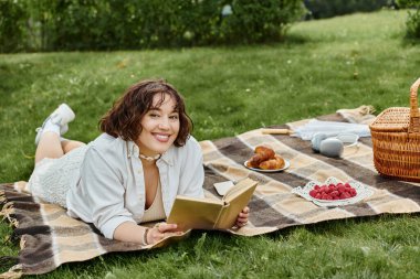 A young woman enjoys a sunny summer day, reading a book while lounging on a picnic blanket filled with treats. clipart