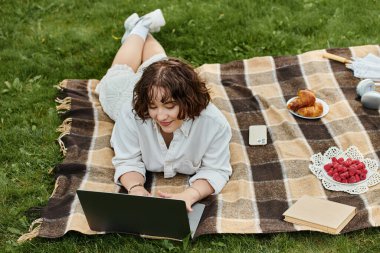 Beyaz tişörtlü genç bir kadın piknik battaniyesinde dinleniyor ve yaz saadeti ortasında laptopuyla çalışıyor..