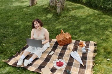 Genç bir kadın piknik battaniyesinde dinleniyor, yaz gününün tadını çıkarırken laptopuyla çalışıyor..