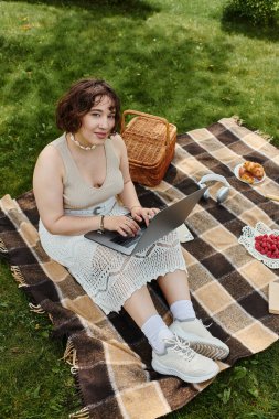 A young woman relaxes in a summer picnic, typing on her laptop while surrounded by delicious treats.