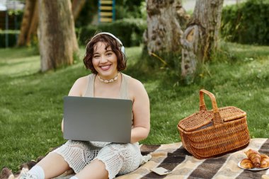 Neşeli bir kadın, yaz sıcağı ve pikniğiyle çevrili, dizüstü bilgisayarına dalmış bir şekilde battaniyeye uzanır..