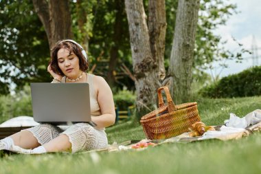Genç bir kadın yaz pikniği sırasında dinleniyor, parkta dizüstü bilgisayarıyla çalışırken müzik dinliyor..