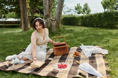 A young woman relaxes on a picnic blanket, enjoying snacks while soaking in the summer sun. clipart