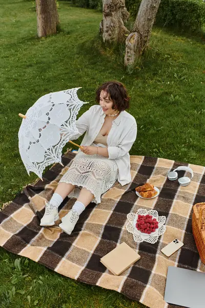Beyaz gömlekli genç bir kadın, bereketli yaz yeşilliğinin ortasında bir şemsiyeyle huzurlu bir pikniğin tadını çıkarıyor..