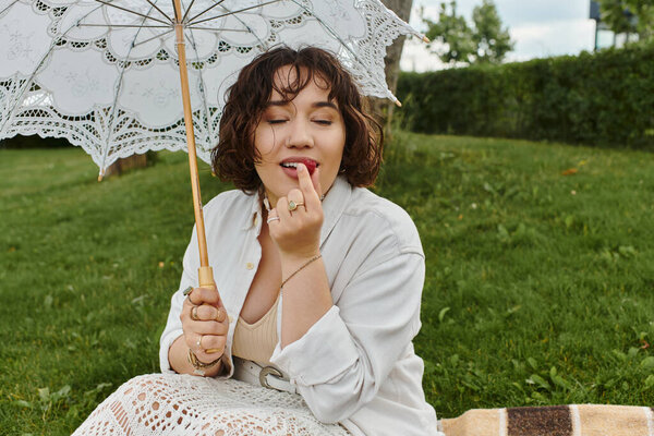 A lovely young woman enjoys a serene moment, applying lip balm under her delicate parasol in the summer sun.