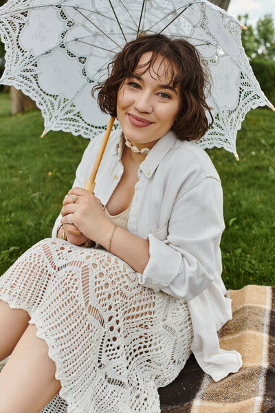 A cheerful young woman relaxes on a blanket, savoring a sunny summer picnic with a delicate umbrella.