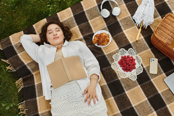 Молодая женщина мирно отдыхает на одеяле для пикника, окруженная вкусной едой под теплым летним солнцем.