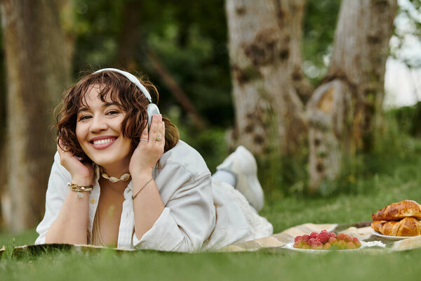 A cheerful young woman relaxes on a blanket, savoring the joys of summer with a delicious picnic spread.