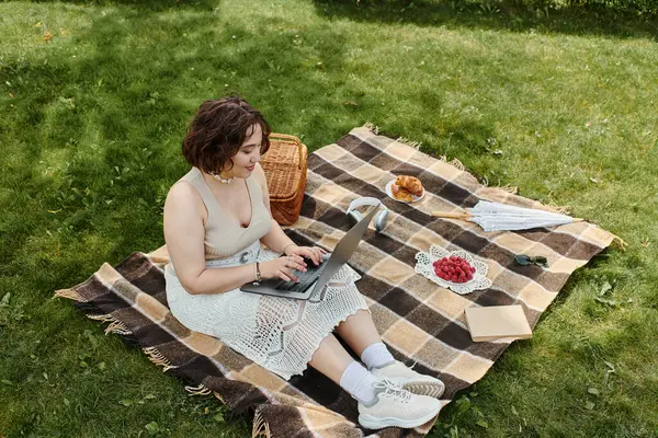 stock image A young woman in a white shirt rests on a blanket, enjoying a summer picnic and working on her laptop.