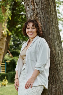 Genç bir kadın canlı bir parkta dinleniyor, yeşilliklerin arasında ılık yaz güneşinin altında ışıl ışıl gülümsüyor..