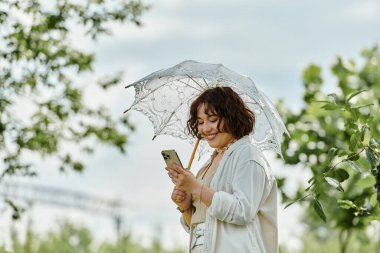 A cheerful woman wears a white shirt, delighting in her phone while shaded by an elegant umbrella in summer greenery. clipart