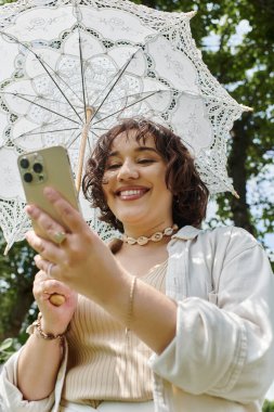 Beyaz giyinmiş neşeli genç bir kadın, telefonuna bakarken yemyeşil bir parkta güneşli bir günün tadını çıkarıyor..