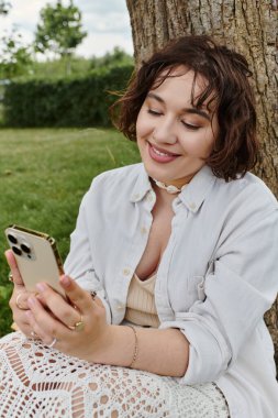 Beyaz tişörtlü neşeli genç bir kadın, güneşli bir parkta telefonuyla meşgulken gülümsüyor..