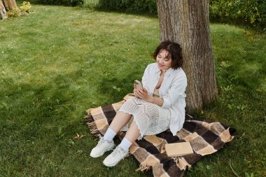 Neşeli genç bir kadın parkta rahat bir battaniyede otururken telefonunu kullanarak dışarıda vakit geçirmekten zevk alıyor..