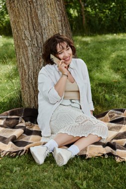 A cheerful young woman in a white shirt relaxes on a blanket in a lush green park during summer sunshine. clipart