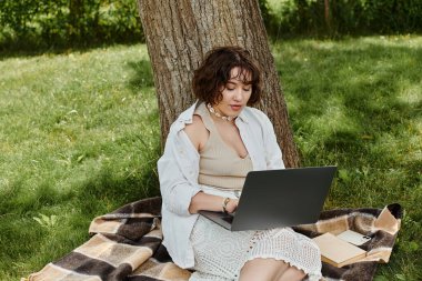 A young woman relaxes in a green park, focused on her laptop, soaking up the summer sun. clipart