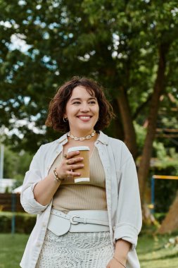 Beyaz gömlekli neşeli genç bir kadın yemyeşil bir parkta güneşi emerken kahvenin tadını çıkarıyor..