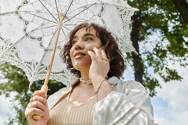 A pretty young woman in a white shirt leisurely chats while holding a lace-covered parasol in a green park.