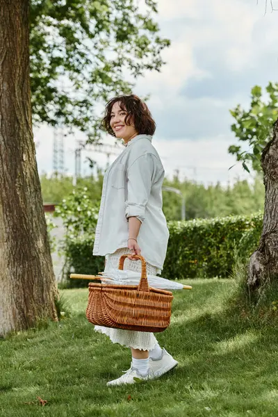 stock image A young woman strolls through a lush green park, carrying a wicker basket under the warm summer sun.