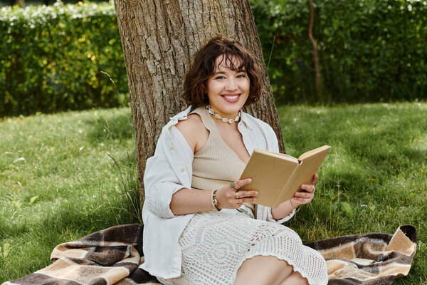 A cheerful young woman enjoys reading a book beneath a shady tree in a vibrant summer park.