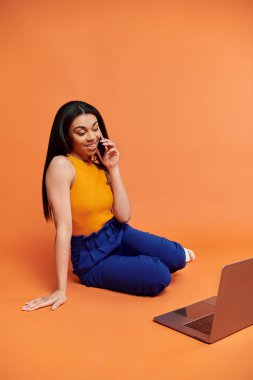 Neşeli bir kadın dizüstü bilgisayarla rahatça otururken telefonla konuşuyor..