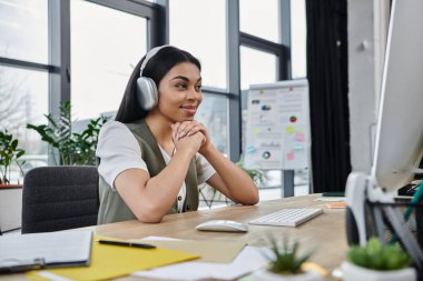 Masasında çalışırken kulaklıklı genç bir kadın gülümsüyor..