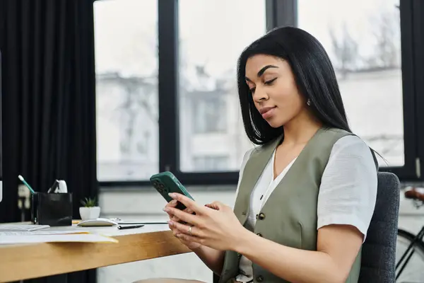 stock image A stylish woman interacts with her smartphone while seated at her desk.